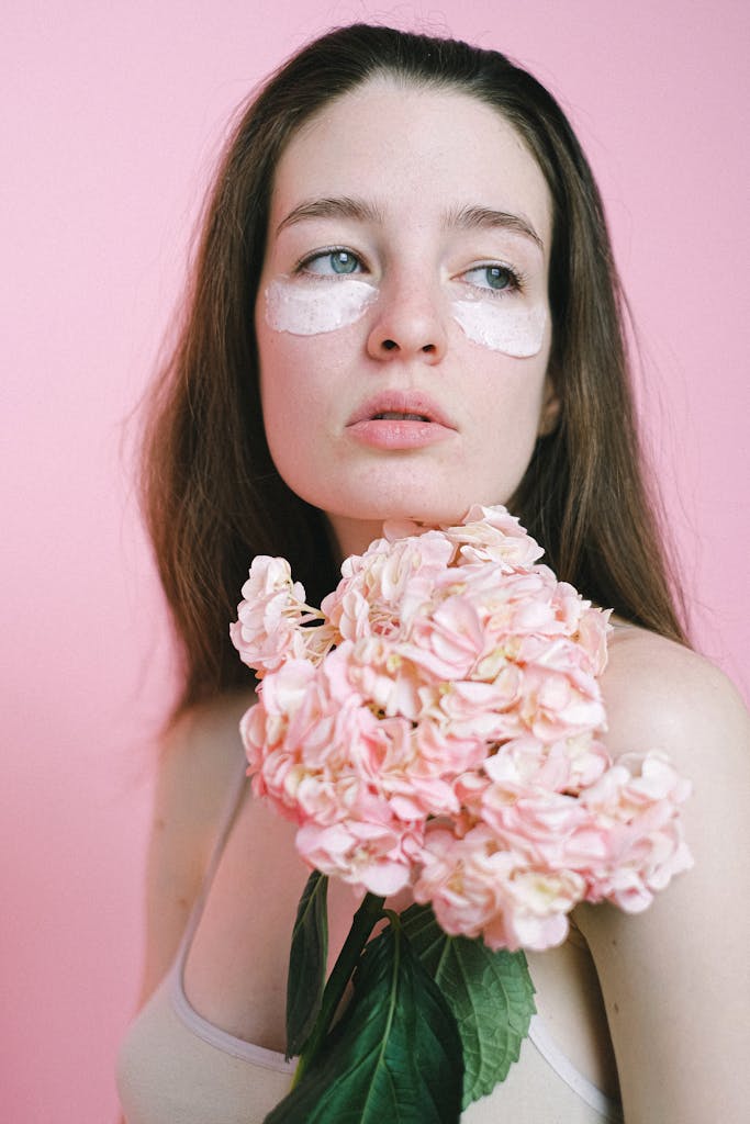 Thoughtful female model with pale skin and anti aging mask under eyes carrying blooming branch of hydrangea while standing against pink background and looking away